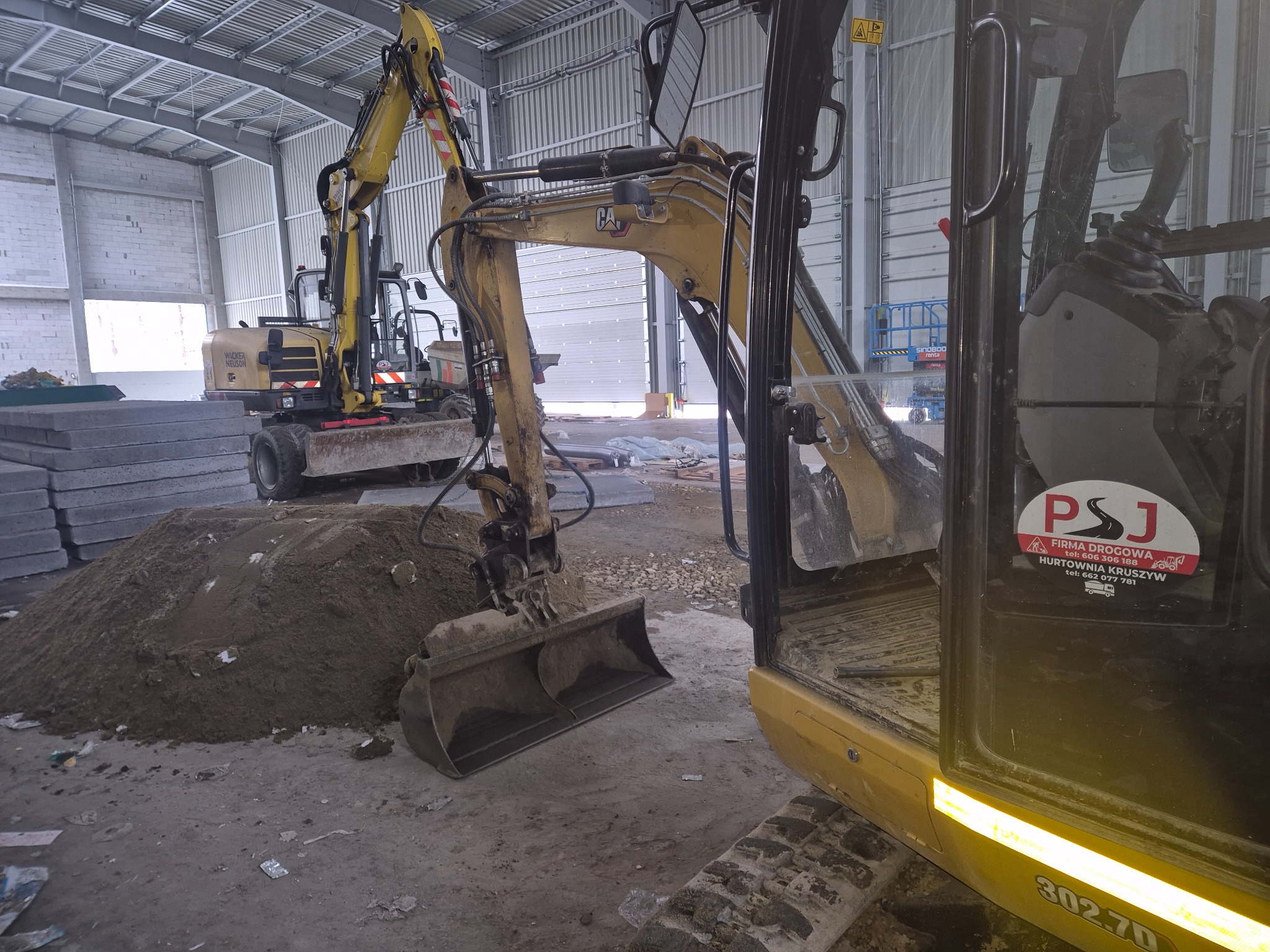 Dwa żółte mini-koparki Caterpillar w hali; jedna z łyżką zagłębia się w pryzmę ziemi, w tle palety z kostką brukową i brama garażowa.