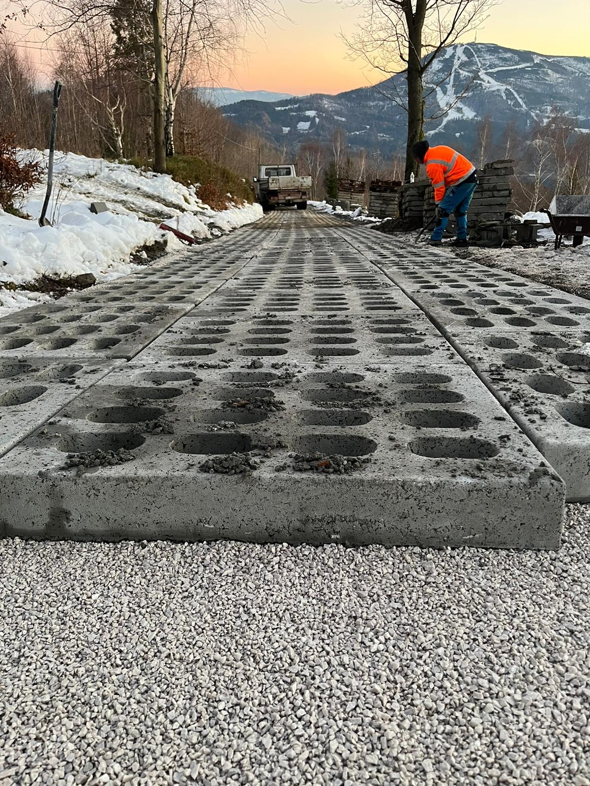 Układanie betonowych płyt ażurowych na podsypce żwirowej w górskim krajobrazie; pracownik w pomarańczowej kurtce wyrównuje teren, a w tle widoczny samochód dostawczy i zalesione wzgórza o zmierzchu.