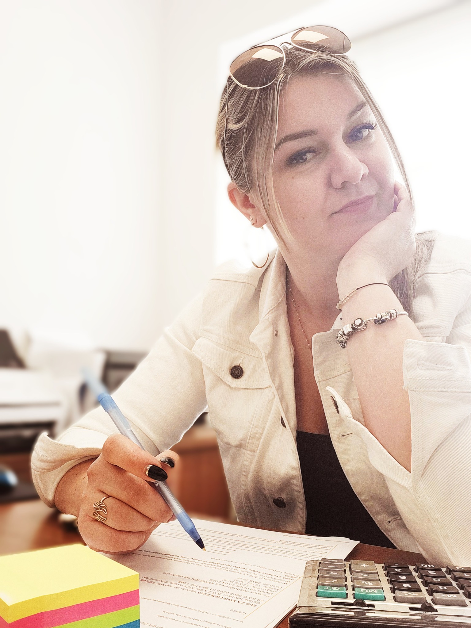 Kobieta w jasnej kurtce pisze na dokumencie, obok kalkulator i karteczki. Uśmiech, okulary na głowie, biżuteria na ręce, ciepłe oświetlenie.