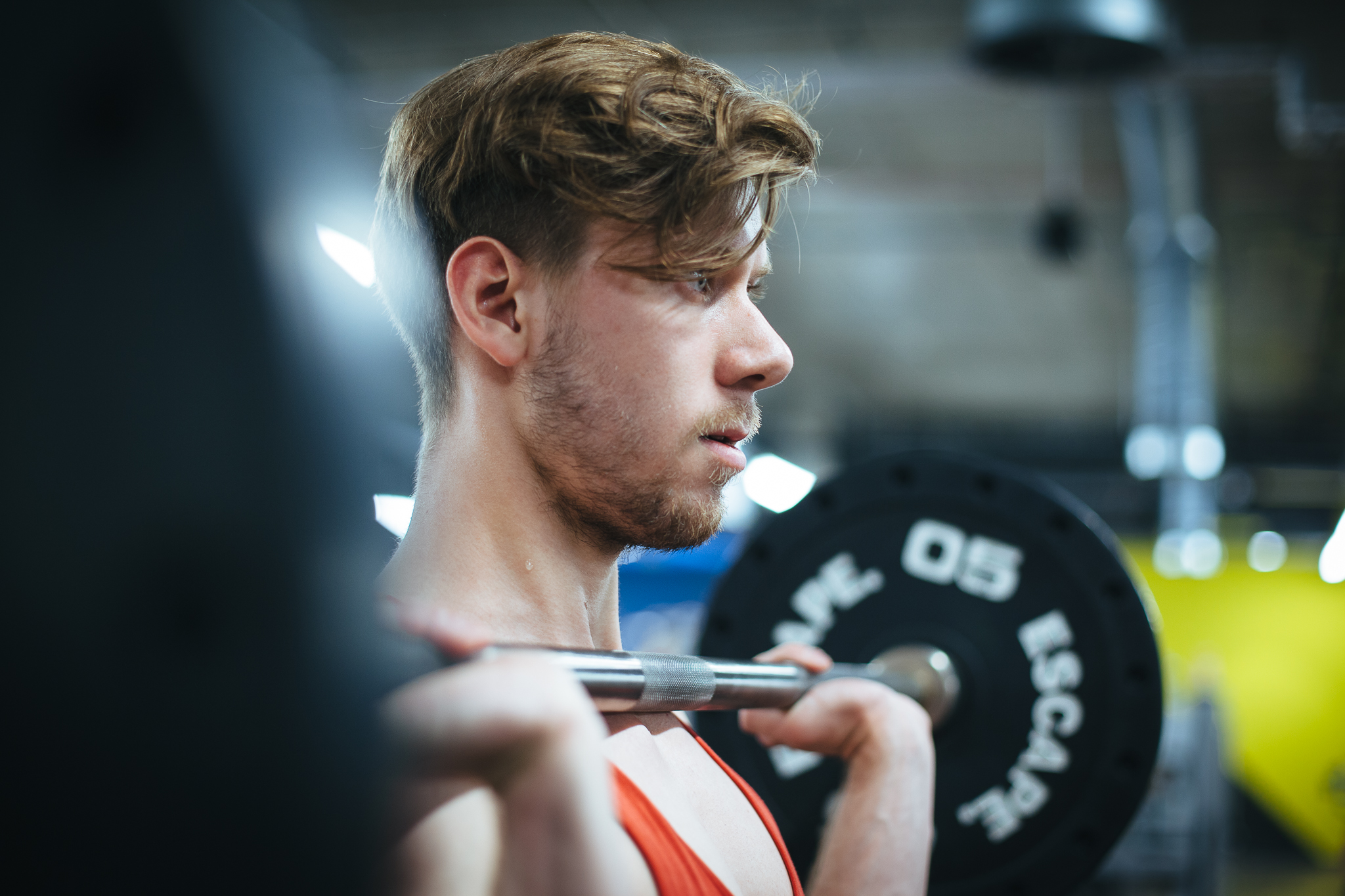 Młody mężczyzna z zarostem, profil twarzy, trzyma sztangę na barkach w siłowni. Widoczne obciążenia na sztandze i fragment wyposażenia siłowni w tle.