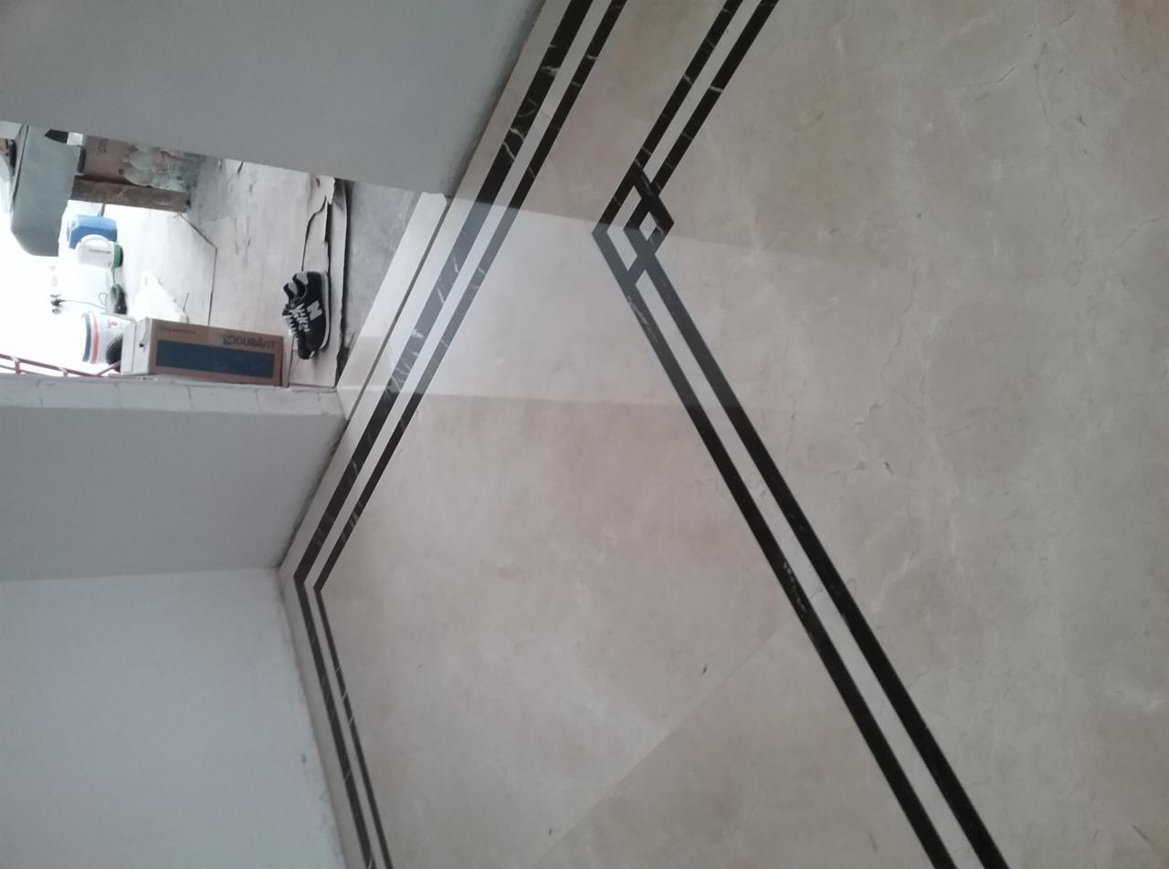 Wykończona podłoga z jasnych płytek z ciemnymi, geometrycznymi wstawkami. W rogu widoczne buty i pudełko z płytkami.