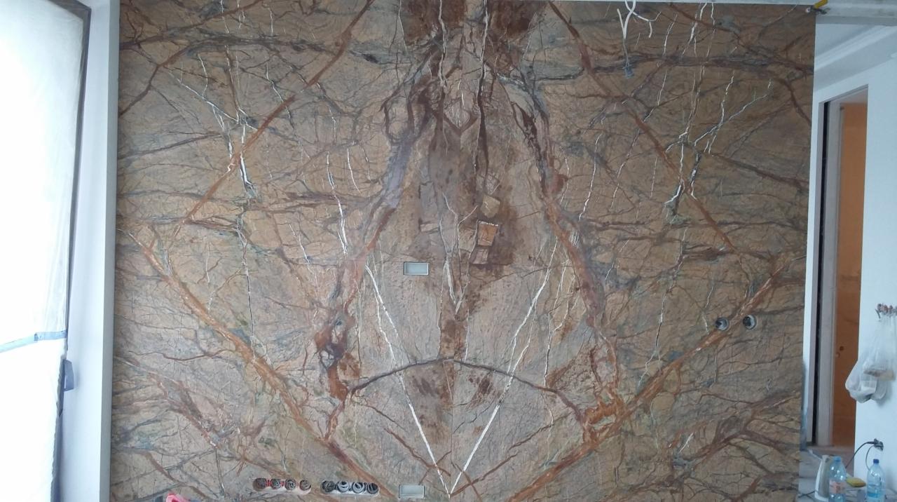 Ściana wyłożona panelami z brązowego kamienia z wyraźnym wzorem żyłkowania, widoczne puszki elektryczne i otwory na kable w ścianie, w tle fragment otwartego przejścia.