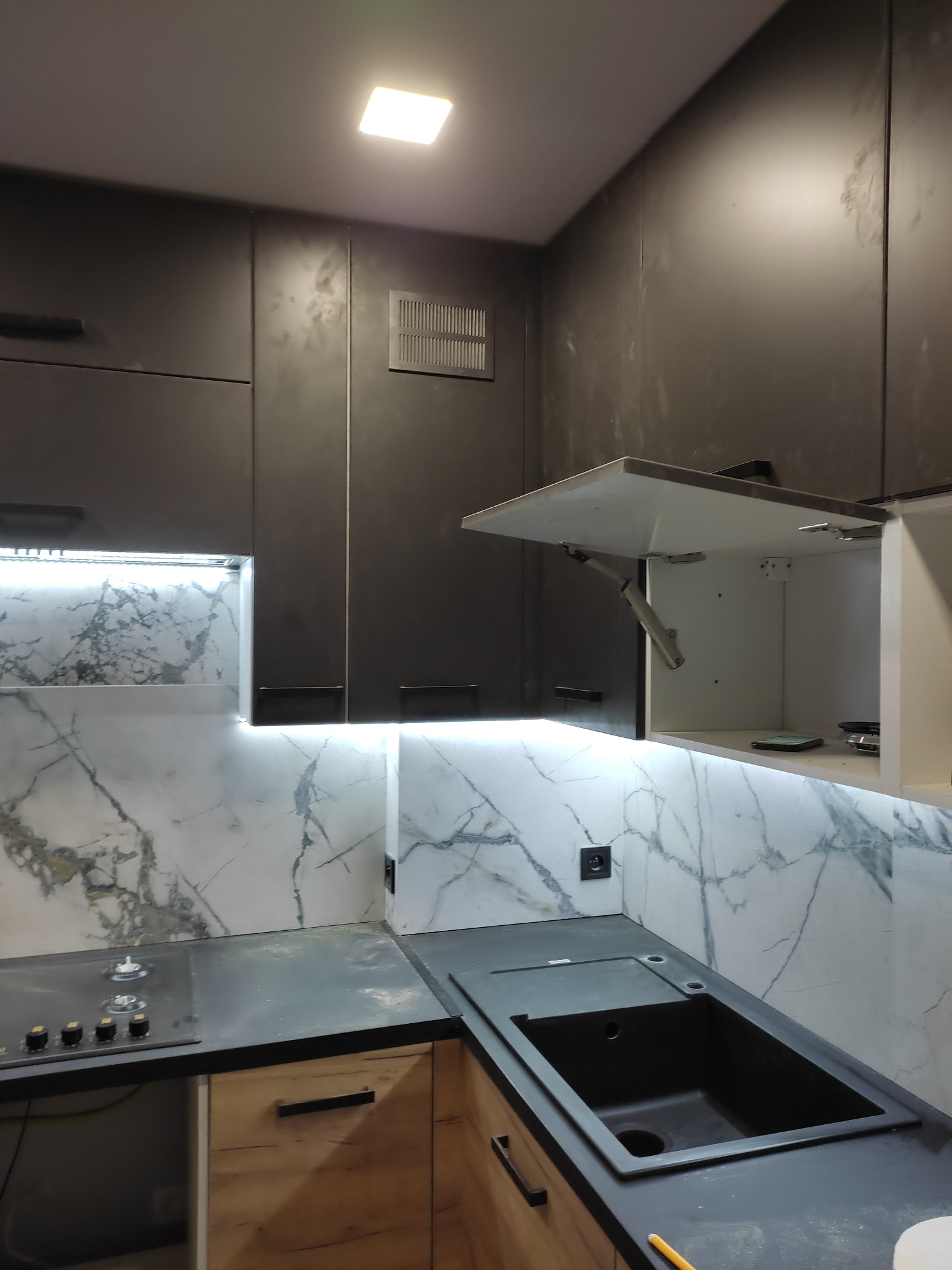 Nowoczesna kuchnia z czarnymi szafkami, marmurowym backsplashiem i oświetleniem LED pod szafkami, z otwartą szafką górną ukazującą mechanizm podnoszenia.
