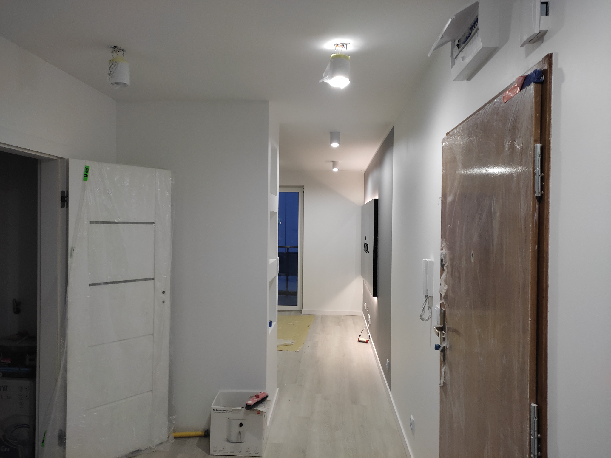 Korytarz w trakcie remontu: zabezpieczone folią drzwi wejściowe i wewnętrzne, widoczne przygotowania do malowania, podłoga częściowo zabezpieczona, oświetlenie sufitowe tymczasowo osłonięte.