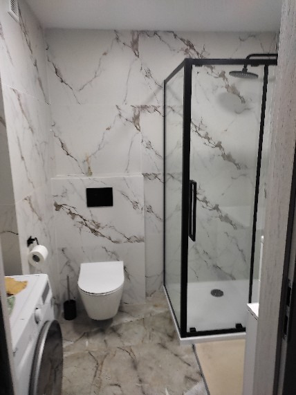Wykończona łazienka z kabiną prysznicową typu walk-in z czarnymi profilami, toaletą podwieszaną, pralką i płytkami imitującymi marmur na ścianach i podłodze.