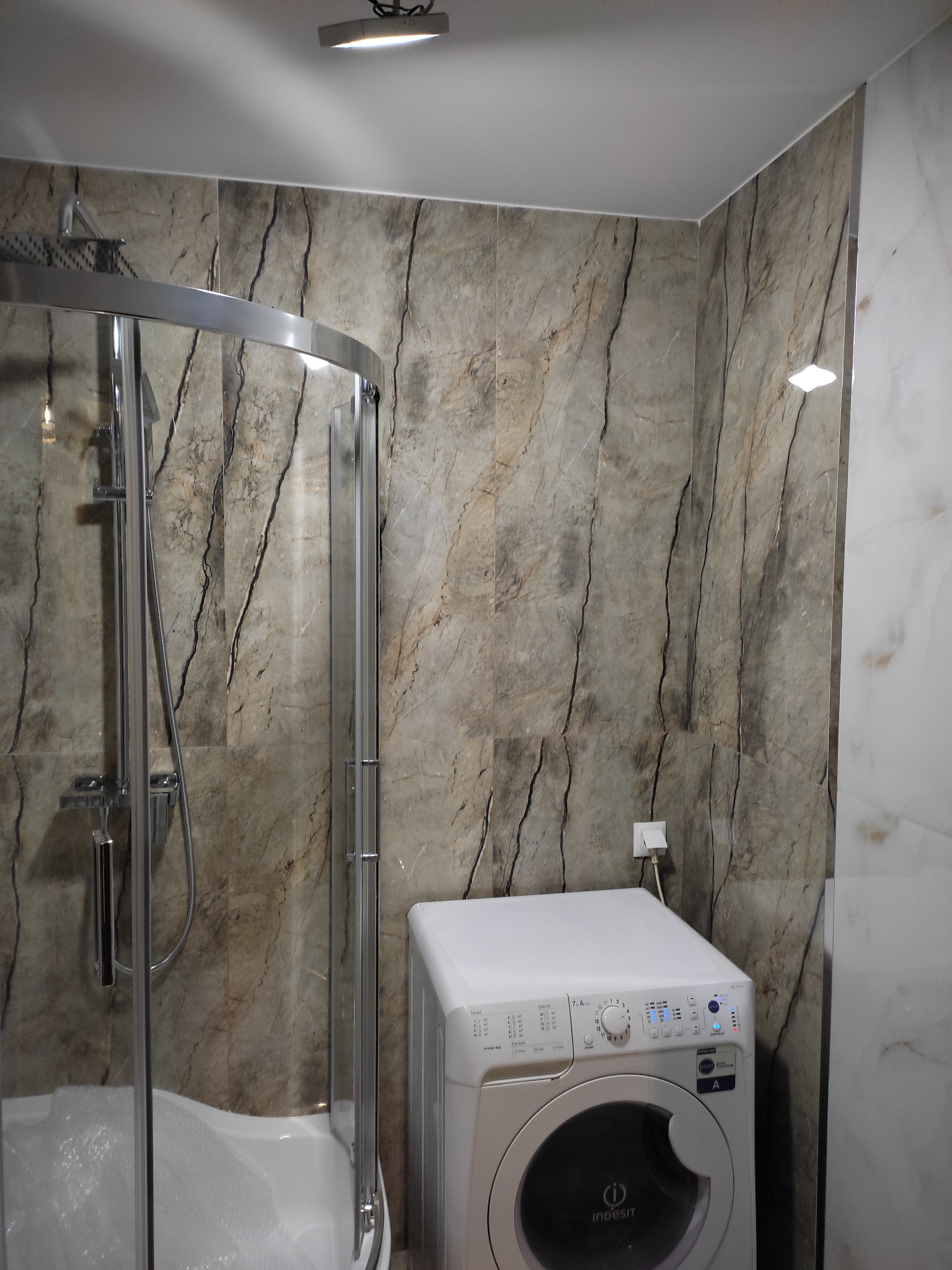Wykończona łazienka z kabiną prysznicową z chromowanymi detalami, marmurowe płytki na ścianach i biała pralka Indesit.