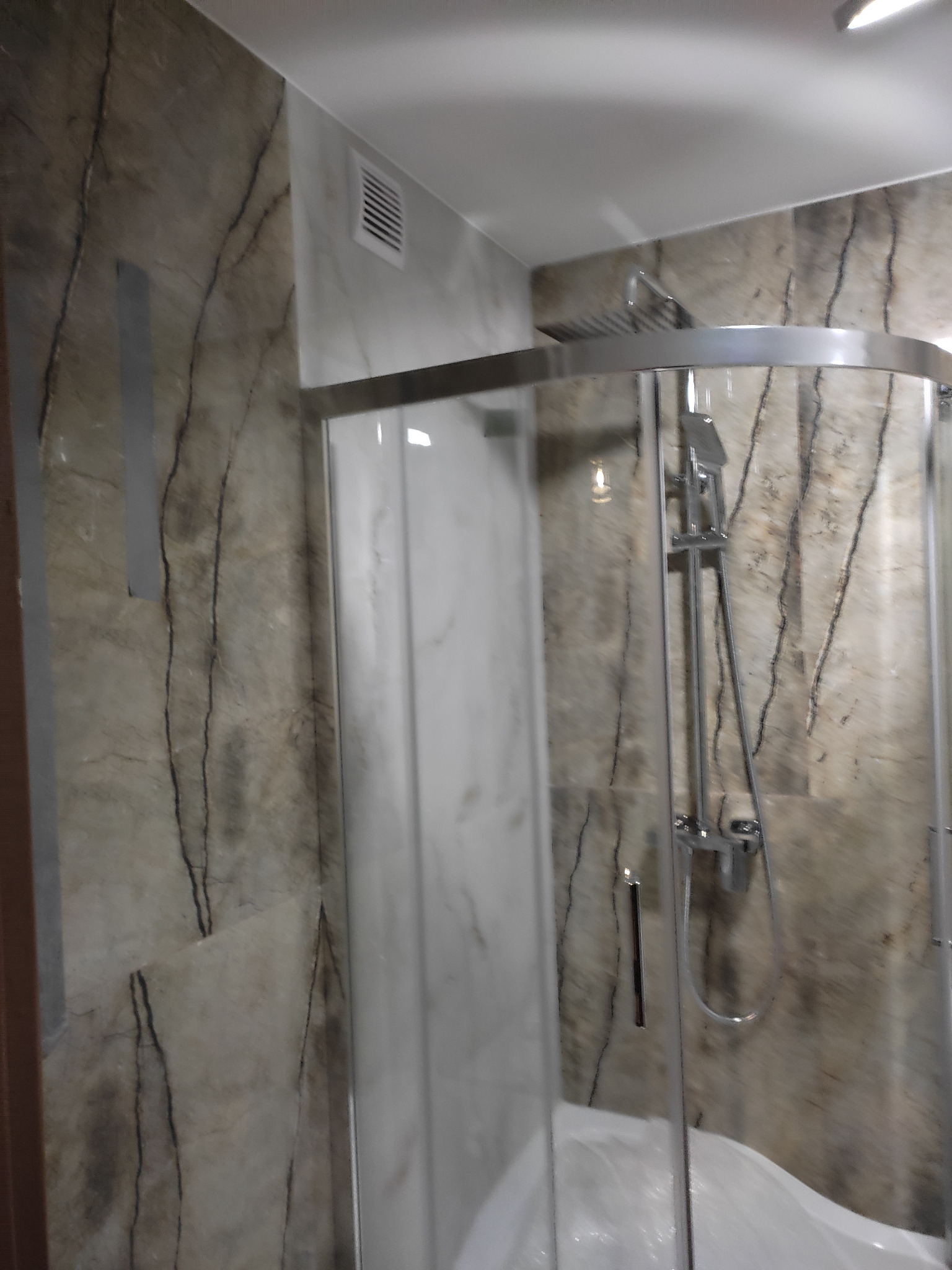 Wykończona kabina prysznicowa z chromowanymi elementami na tle ściany wyłożonej płytkami imitującymi marmur z wyraźnym rysunkiem żyłkowania.