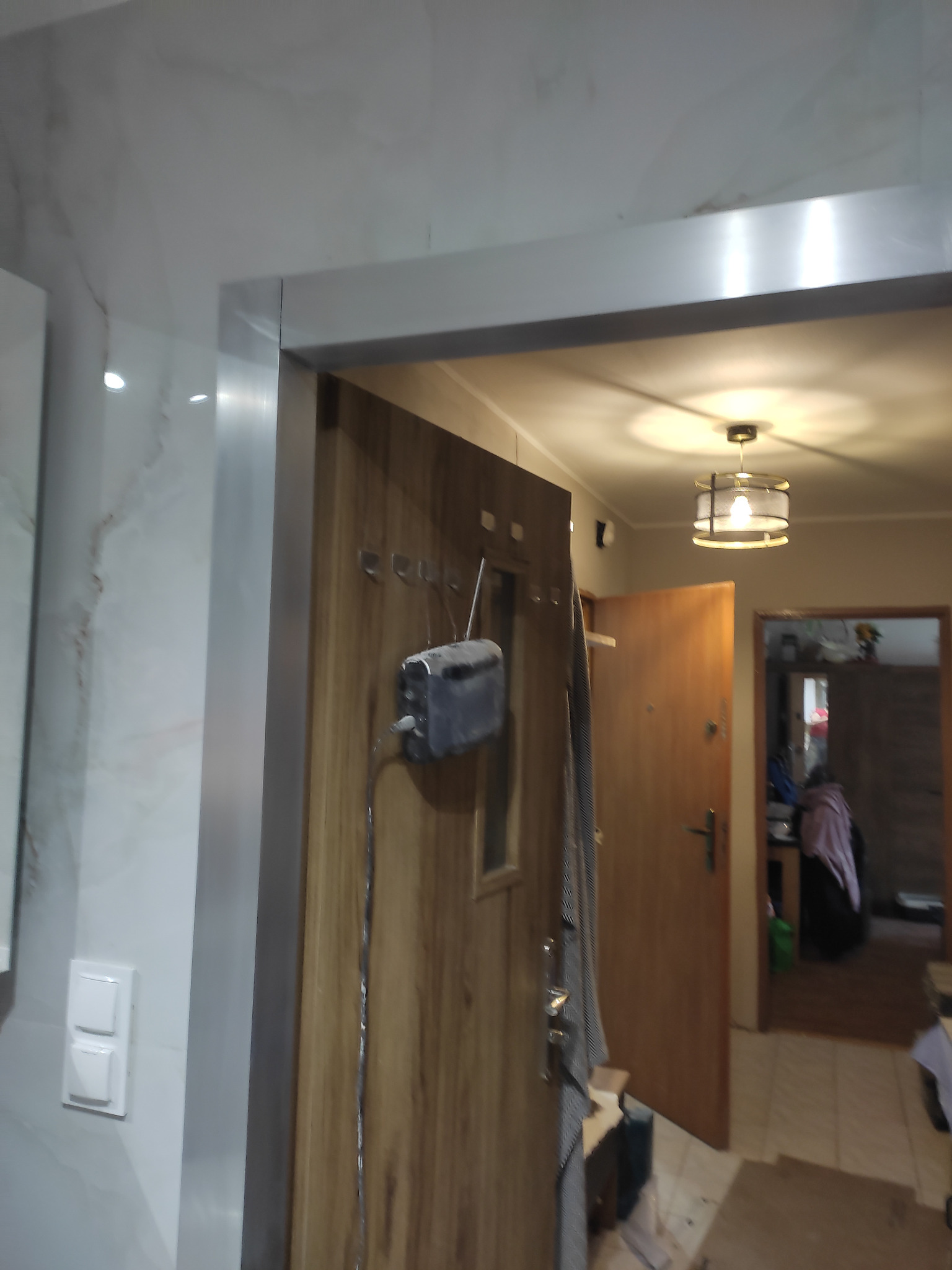 Wykończona łazienka z marmurowymi płytkami i drewnianymi drzwiami. Widoczny włącznik światła i lampa sufitowa. W tle fragment wnętrza mieszkania.