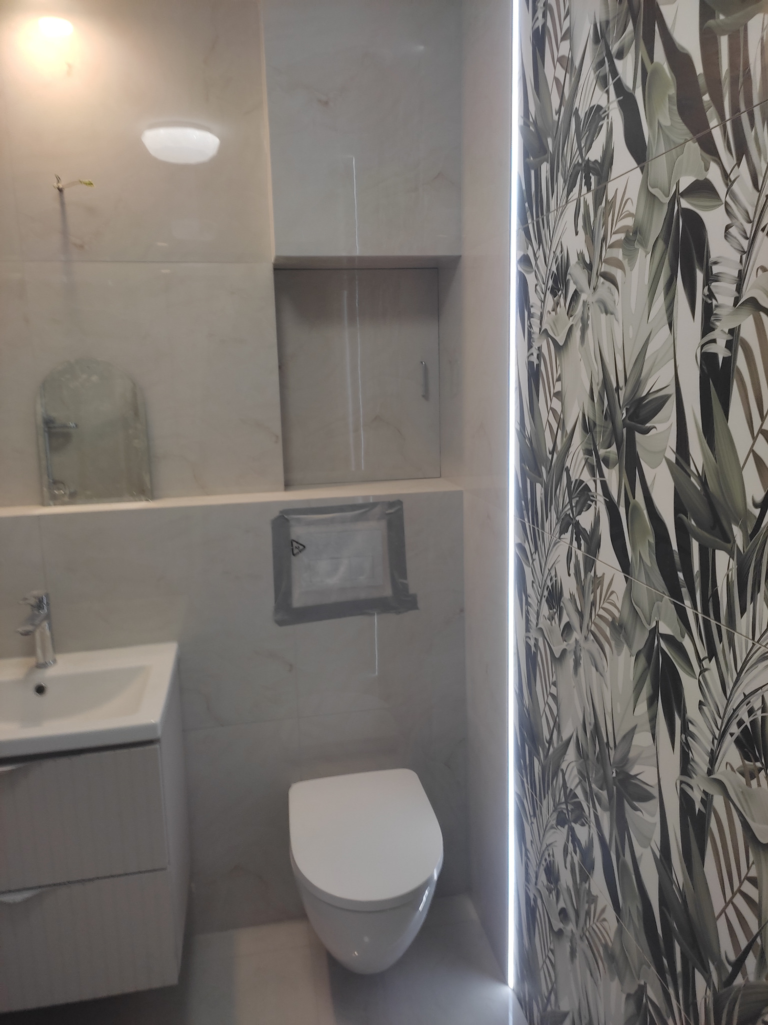 Wykończona łazienka z umywalką, toaletą i płytkami w tropikalne wzory. Widoczne oświetlenie LED i ukryte schowki. Minimalistyczny design.