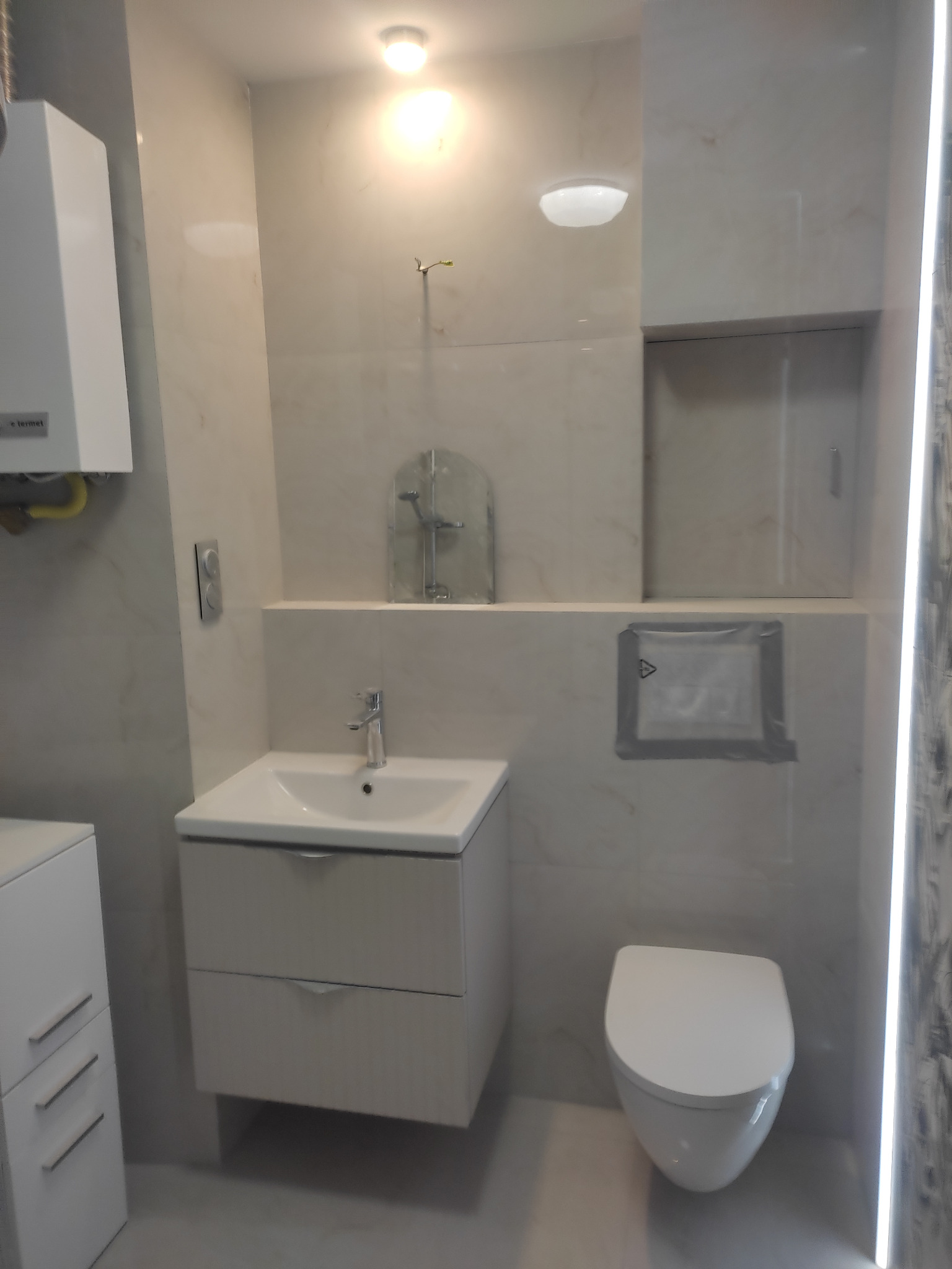 Jasna łazienka z umywalką na szafce, toaletą i grzejnikiem. Ściany wyłożone płytkami imitującymi marmur. Widoczne oświetlenie LED.