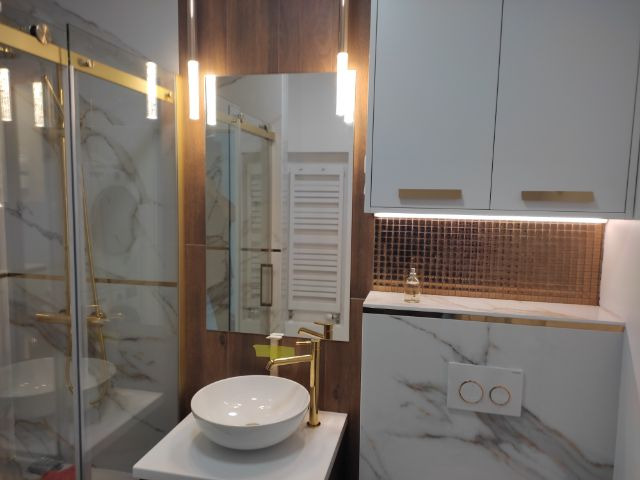 Elegancka łazienka z kabiną prysznicową, umywalką nablatową i złotymi akcentami. Białe marmurowe płytki, drewniane detale i oświetlenie LED tworzą luksusowy klimat.