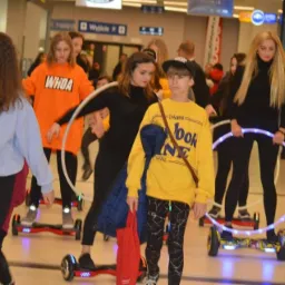 Grupa nastolatków na hoverboardach z hula-hoop w hali, niektóre z hula-hoop świecą, w tle widoczni przechodnie i tablice informacyjne.