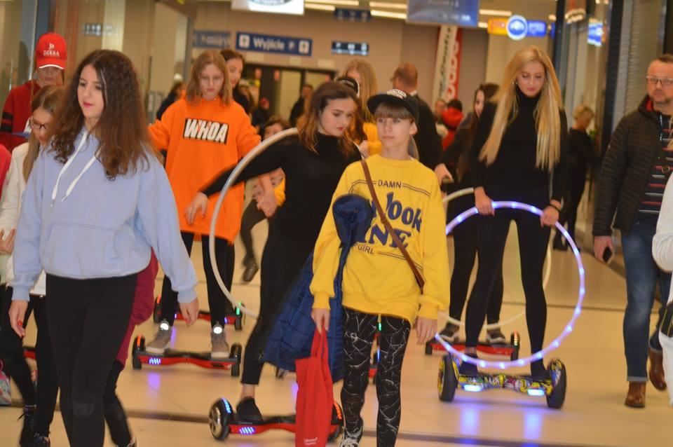 Grupa nastolatków na hoverboardach z hula-hoop w hali, niektóre z hula-hoop świecą, w tle widoczni przechodnie i tablice informacyjne.