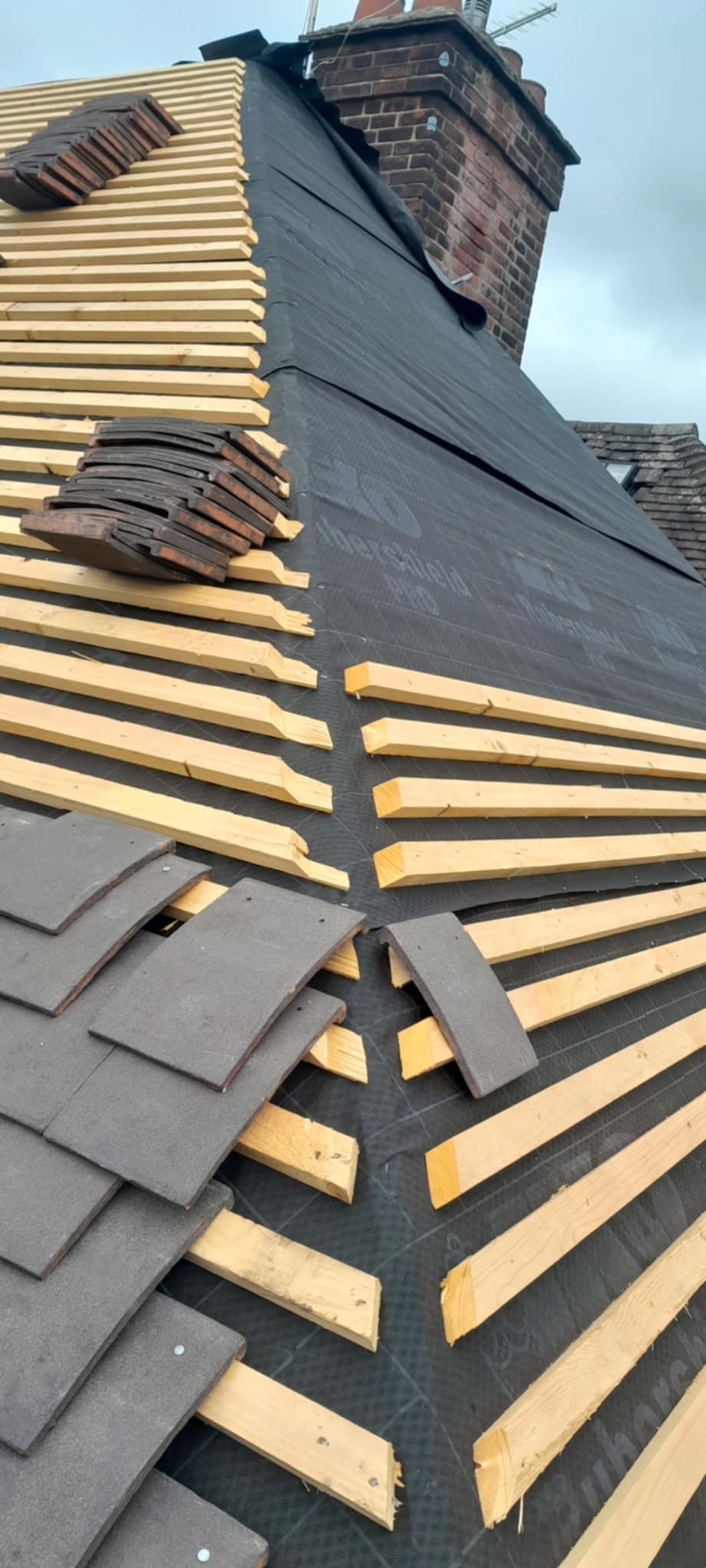 Montaż łat pod dachówkę na czarnej membranie dachowej. Częściowo ułożona dachówka ceramiczna. Widoczny komin z cegły. Prace dekarskie w toku.