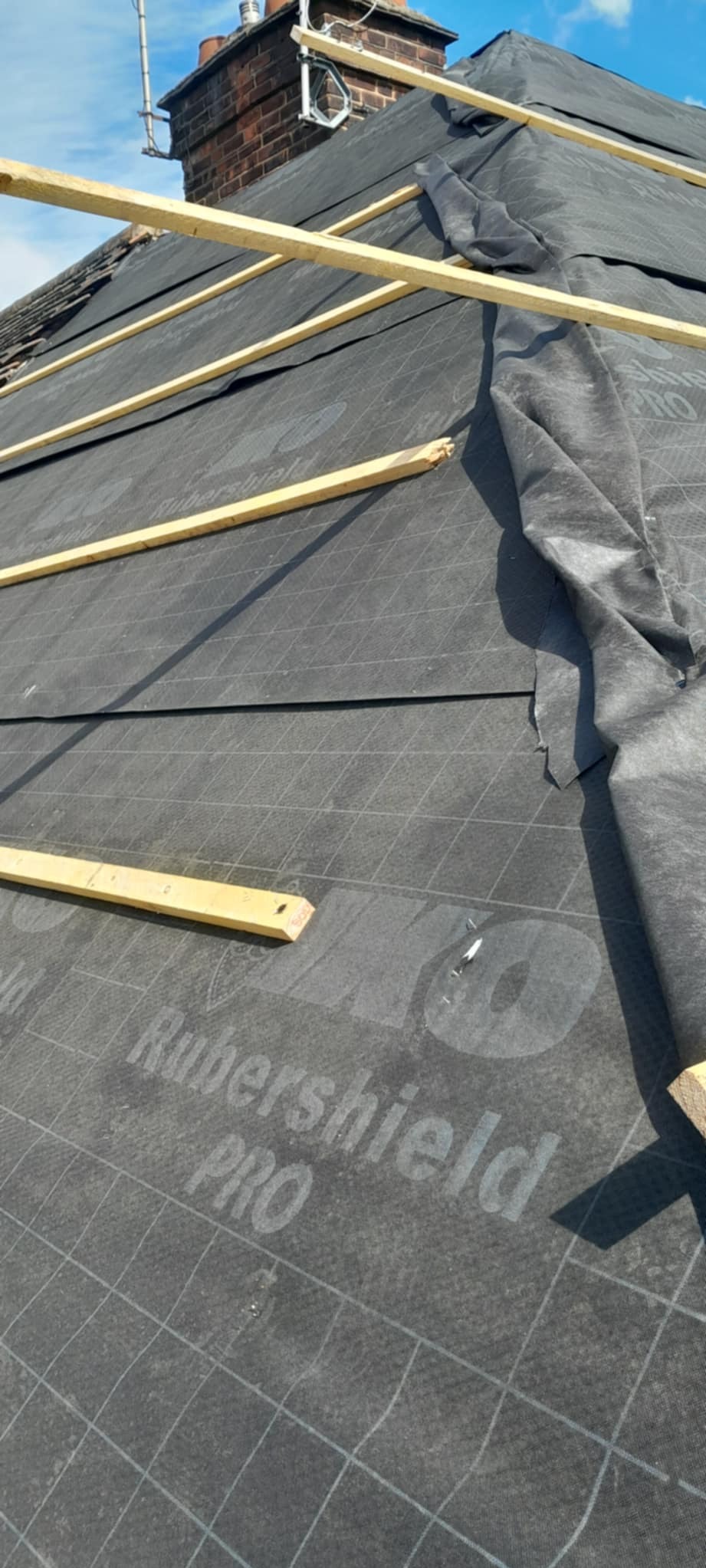 Czarny materiał hydroizolacyjny z napisem Rubershield PRO, przytwierdzony do dachu drewnianymi łatami. W tle komin i fragment nieba.