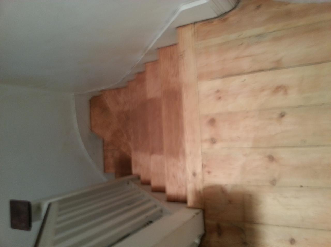 Wnętrze w trakcie wykańczania: surowe drewniane schody z widocznym grzejnikiem, ściany częściowo wykończone boazerią, sufit pomalowany na biało.