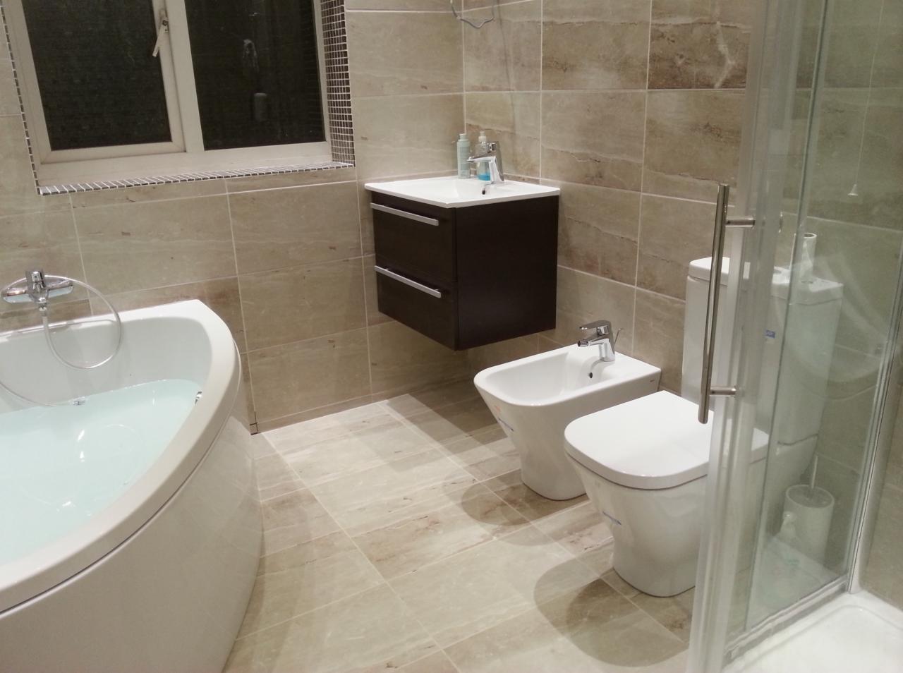 Nowoczesna łazienka z beżowymi płytkami imitującymi marmur, wyposażona w białą wannę narożną wypełnioną wodą, podwieszaną szafkę z umywalką, bidet, toaletę i kabinę prysznicową ze szklanymi drzwiami.