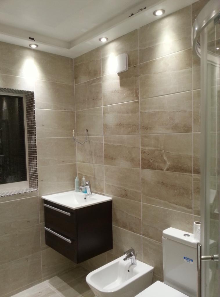 Nowoczesna łazienka z beżowymi płytkami imitującymi marmur, podwieszana szafka z umywalką, bidet i toaleta w komplecie, oświetlenie punktowe w suficie.