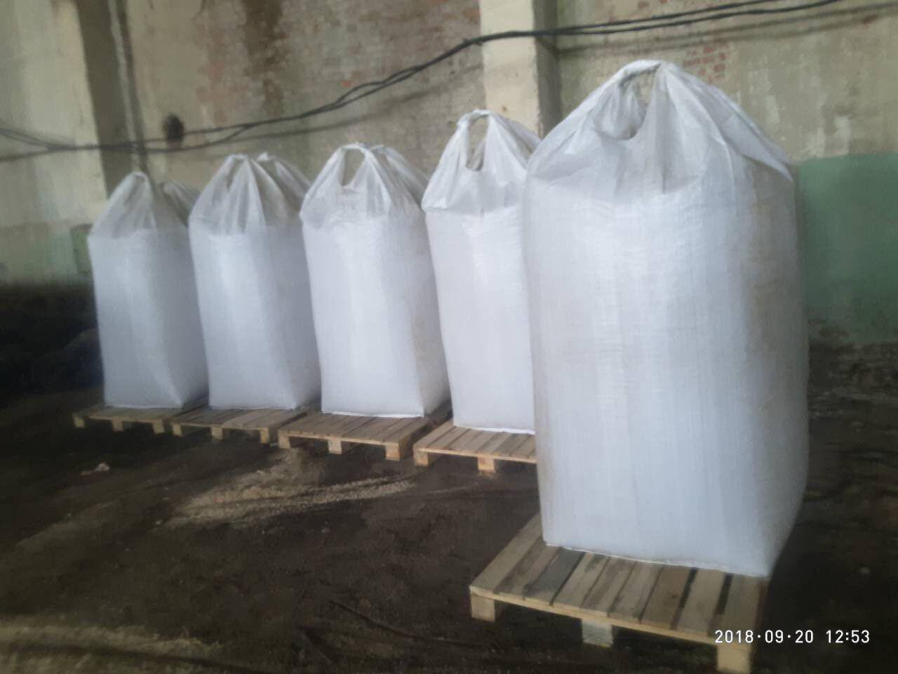 Sześć dużych, białych worków z pelletem ustawionych w rzędzie na drewnianych paletach w magazynie z betonowymi ścianami i podłogą pokrytą trocinami.