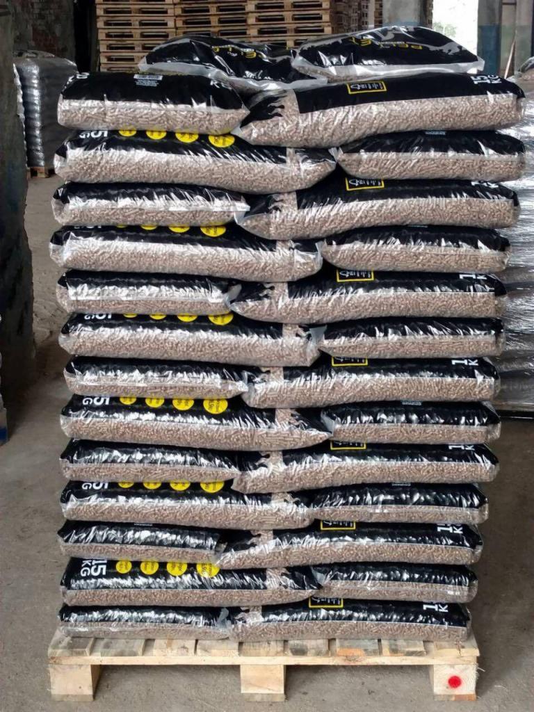 Paleta z ułożonymi workami z pelletem drzewnym, czarne worki z widoczną zawartością, żółte naklejki na workach.