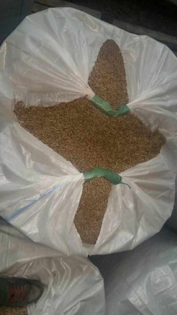 Otwarty worek typu big bag wypełniony jasnobrązowym granulatem, zamknięty dwoma zielonymi zaciskami, widoczny fragment buta w lewym dolnym rogu.