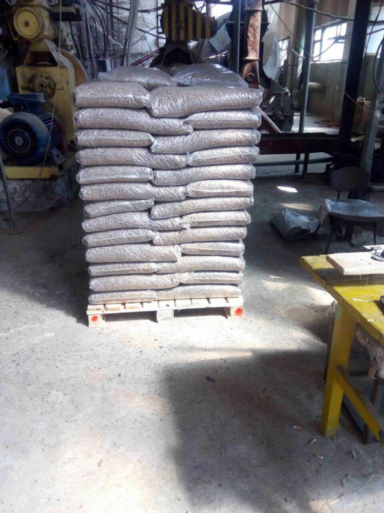 Stos worków z granulatem drzewnym ułożonych na palecie w hali produkcyjnej, widoczny fragment maszyny i żółtego stołu.