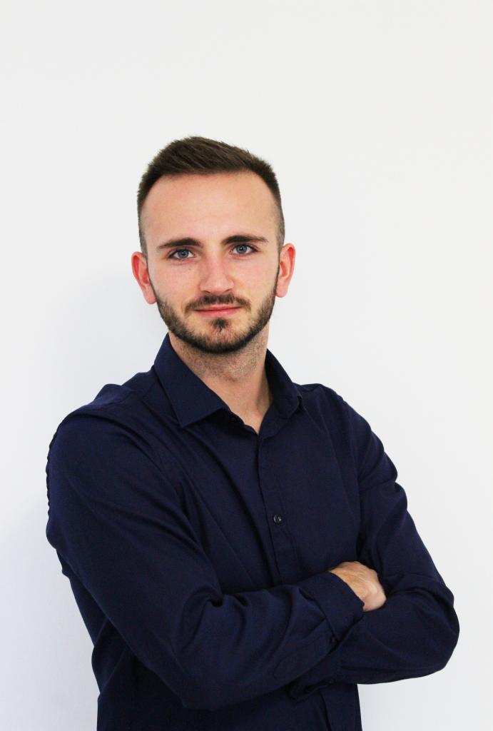 Młody mężczyzna w granatowej koszuli, z lekkim zarostem i niebieskimi oczami, stoi na białym tle z założonymi rękami.