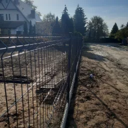 Nowo postawione, ciemne ogrodzenie panelowe z widocznymi betonowymi fundamentami, biegnące wzdłuż piaszczystej drogi gruntowej, w tle dom jednorodzinny i drzewa, w słoneczny dzień.