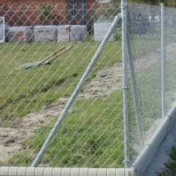 Fragment ogrodzenia z siatki z metalowymi słupkami i betonową podmurówką na trawiastym terenie, w tle ceglany budynek i materiały budowlane.