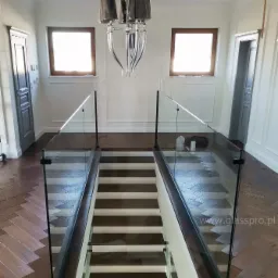 Nowoczesne szklane balustrady zabezpieczające schody w jasnym wnętrzu z parkietem w jodełkę i eleganckim żyrandolem.