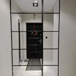 Szklana ścianka działowa z czarnymi szprosami, widok na korytarz z czarnymi drzwiami wejściowymi ozdobionymi świątecznym wieńcem, marmurowa podłoga.