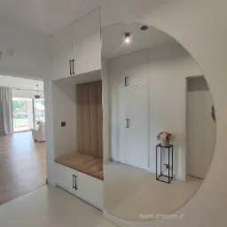 Nowoczesny przedpokój z zabudową meblową: białe szafy z czarnymi uchwytami, siedzisko z tapicerowanym oparciem w odcieniach beżu oraz duże okrągłe lustro z widocznym odbiciem wnętrza.