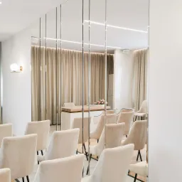 Jasne krzesła z welurową tapicerką ustawione rzędami w sali z lustrzanymi panelami ściennymi, widoczne oświetlenie LED i minimalistyczny kinkiet.