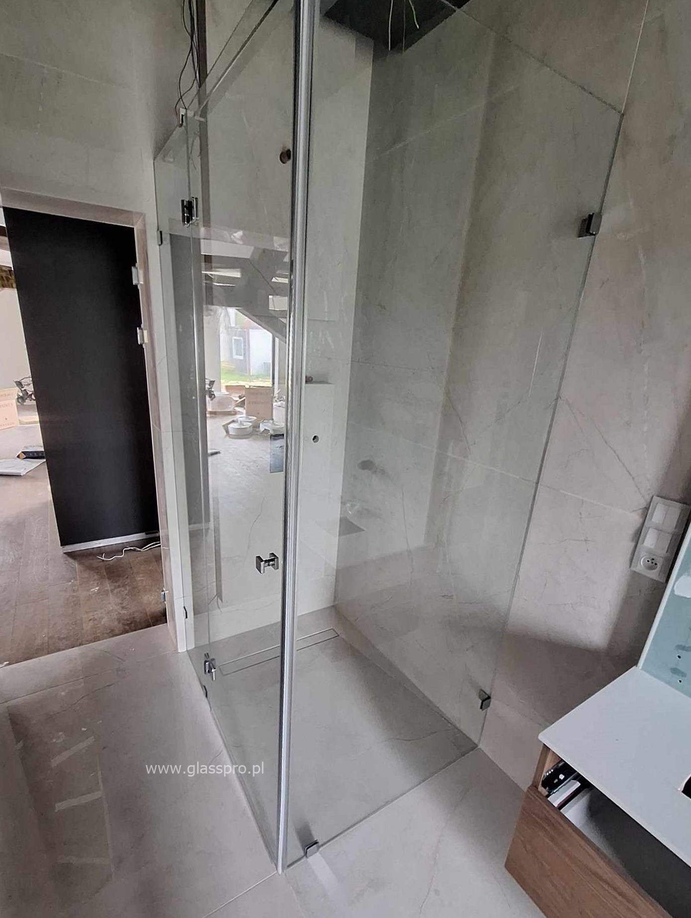 Nowoczesna kabina prysznicowa ze szkła hartowanego z minimalistycznymi okuciami, widoczna w surowym wnętrzu łazienki w trakcie wykończenia, z widocznymi przewodami i elementami instalacji.
