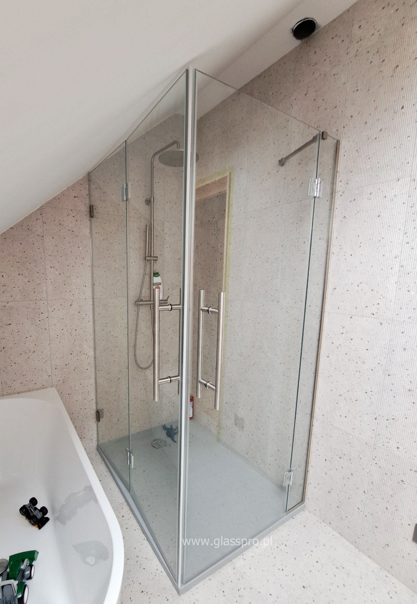 Kabina prysznicowa ze szkła hartowanego w łazience na poddaszu, z widocznymi zawiasami i uchwytami ze stali nierdzewnej, obok wanna z zabawkami.