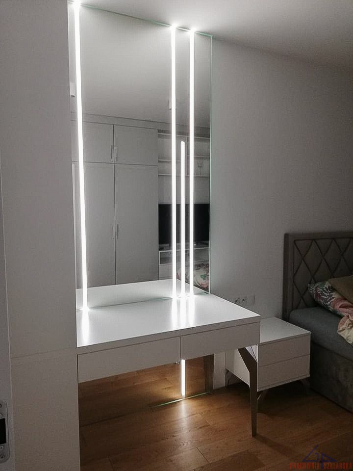 Biała toaletka z lustrem, podświetlonym pionowymi paskami LED, odbijająca wnętrze szafy w minimalistycznej sypialni z drewnianą podłogą.