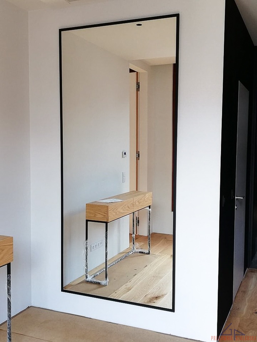 Duże lustro w czarnej ramie, odbijające konsolę z jasnego drewna na metalowych nogach, zabezpieczonych folią ochronną, w minimalistycznym wnętrzu z drewnianą podłogą.