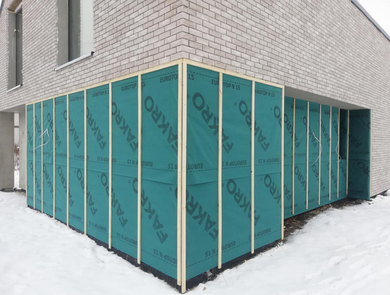 Etap budowy domu murowanego: montaż izolacji przeciwwilgociowej FAKRO EUROTOP N 15 na drewnianym stelażu, widoczny fragment ściany z jasnej cegły i śnieg na ziemi.