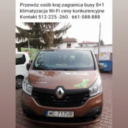Brązowy bus Renault z naklejką psa rasy jamnik na masce, tablica rejestracyjna WE 717UR, na tle budynków i drzew. Reklama przewozu osób.