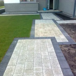 Ścieżka z jasnej kostki z ciemną obwódką prowadząca do wejścia domu, obok trawnik i rabata.