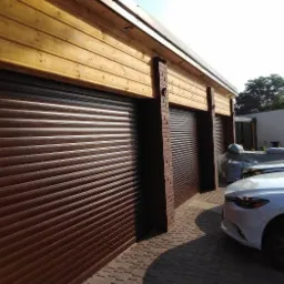 Zewnętrzny widok garażu z trzema brązowymi rolowanymi bramami, wykończonego drewnianą elewacją i ceglanymi filarami, z fragmentem białego samochodu w tle.