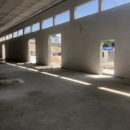 Surowe wnętrze budynku w trakcie prac wykończeniowych: świeżo otynkowane ściany, betonowa podłoga z resztkami materiałów budowlanych, promienie słoneczne wpadające przez otwory okienne i drzwiowe.