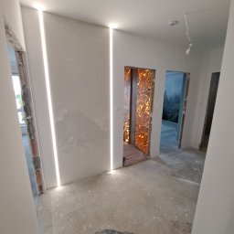 Mochocki Piotr - Wnętrze w trakcie wykańczania: surowe ściany z oświetleniem LED, dekoracyjna okładzina wnęki drzwiowej, surowa podłoga. Widoczne przygotowania do dalszych prac.