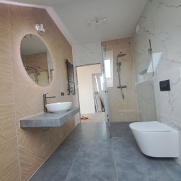 Mochocki Piotr - Nowoczesna łazienka z beżowymi i białymi płytkami, szarą podłogą, owalnym lustrem i minimalistyczną armaturą. Widoczny fragment kabiny prysznicowej i toalety.