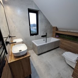 Mochocki Piotr - Nowoczesna łazienka na poddaszu z dwoma umywalkami na drewnianym blacie, wanną i oknem. Ściany wyłożone szarymi płytkami, drewniane lamele dekoracyjne.