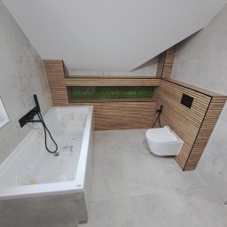 Mochocki Piotr - Nowoczesna łazienka na poddaszu z wanną w folii, wiszącą toaletą i drewnianą zabudową z zielonym akcentem. Szare płytki na ścianach i podłodze.