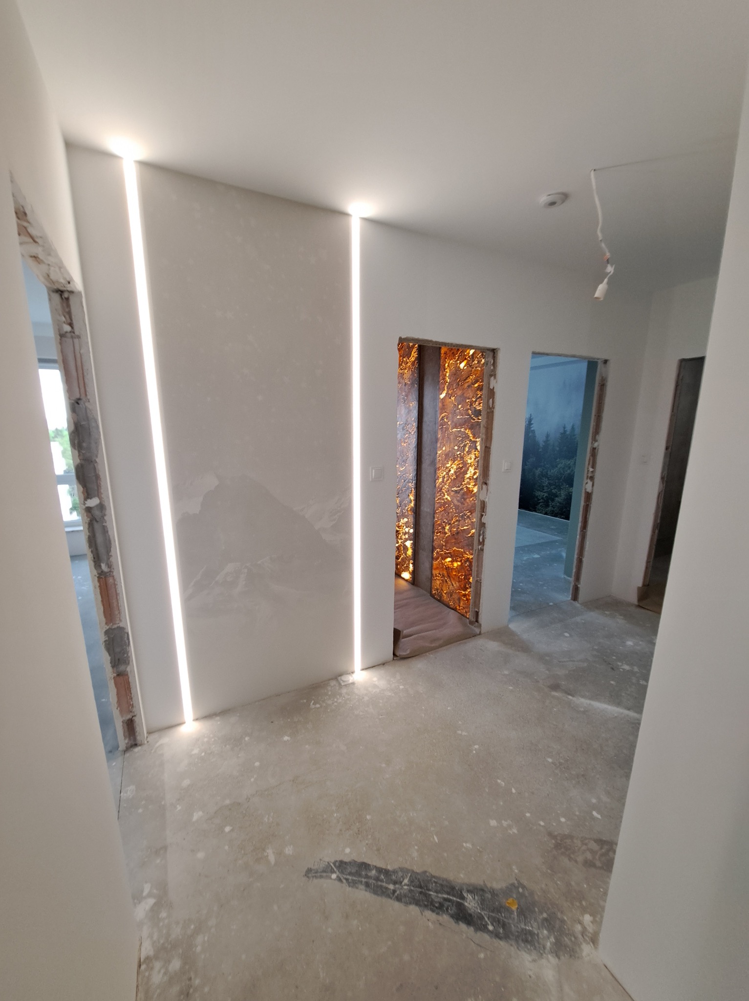 Wnętrze w trakcie wykańczania: surowe ściany z oświetleniem LED, dekoracyjna okładzina wnęki drzwiowej, surowa podłoga. Widoczne przygotowania do dalszych prac.