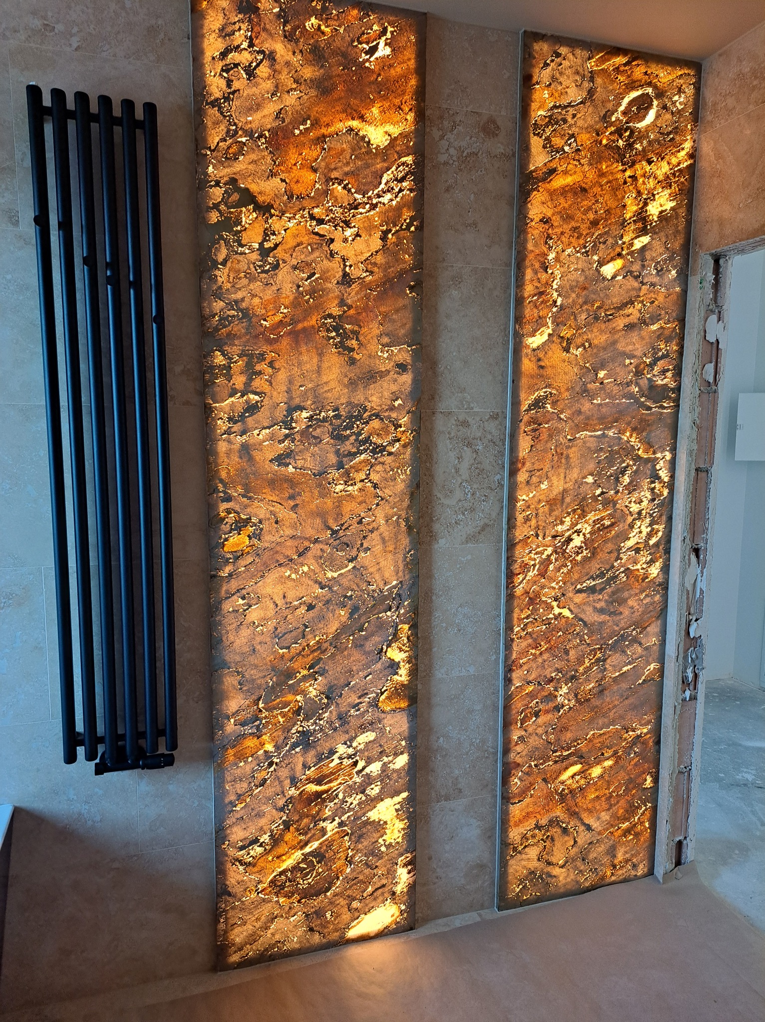 Podświetlane panele dekoracyjne z efektem kamienia w łazience, obok czarny grzejnik drabinkowy. Widoczne płytki na ścianach i surowy stan wykończenia przy drzwiach.