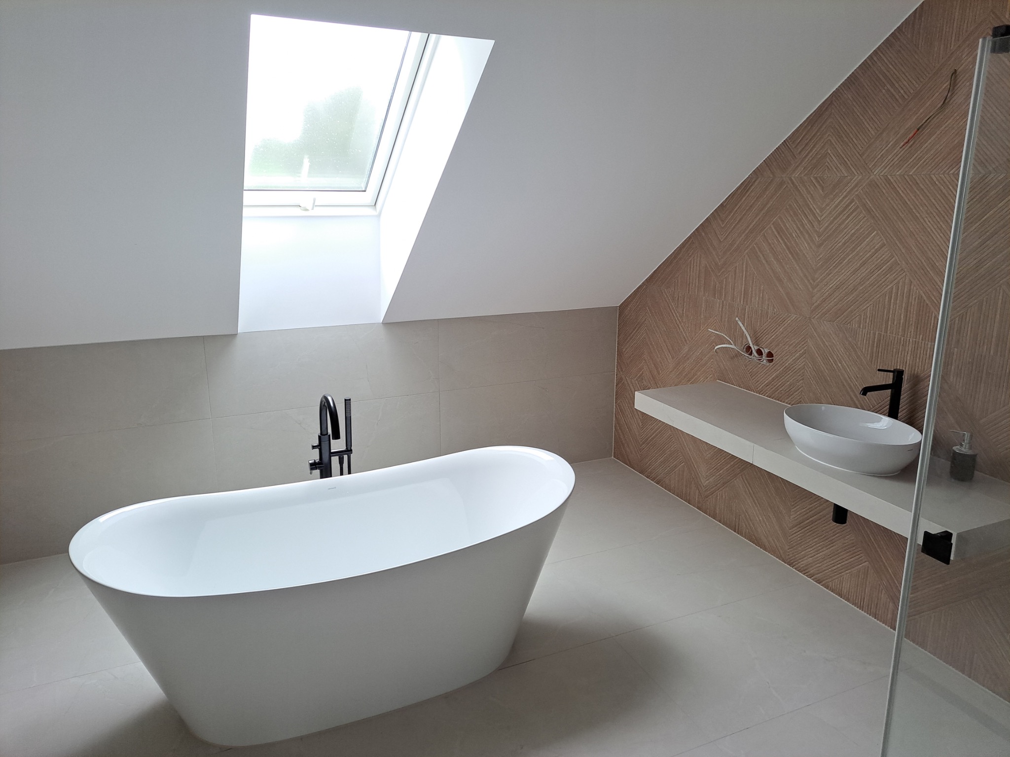 Nowoczesna łazienka na poddaszu z wolnostojącą wanną, umywalką na blacie i oknem dachowym. Beżowe płytki i geometryczny wzór na ścianie dodają elegancji.