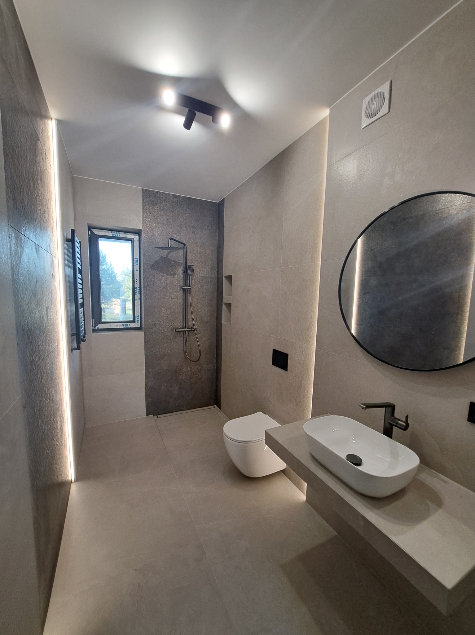 Nowoczesna łazienka w beżach i szarościach z oświetleniem LED, okrągłym lustrem i wiszącą toaletą. Minimalistyczny design z akcentami czerni.